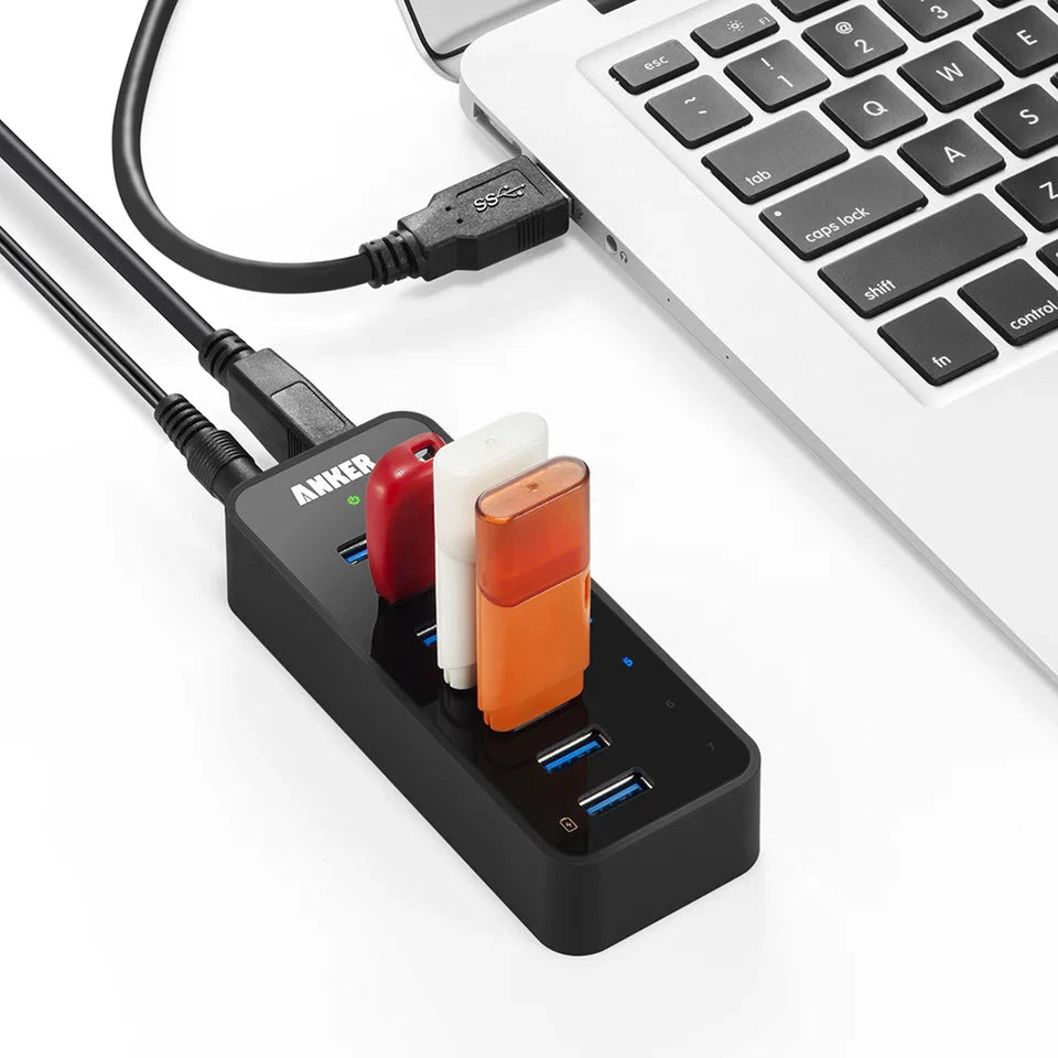 Anker 7-Port USB 3.0 hub with charging port, black, connected to a laptop with three colored USB devices plugged in.